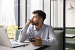 © fizkes - Pensive, thoughtful young male freelancer sit at desk with laptop in modern coworking office holding phone waiting for response, reviewing received information, or considering work or personal matter