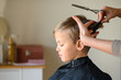 © Mariia Andreeva - A little blond boy gets a haircut, sitting in a salon chair with a cape. The hairdresser holds scissors and a comb. The child is calm and enjoying the experience. Concept of childhood grooming and sel