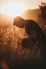 Poster - A man is seen kneeling in a vast field of tall grass, with the sun shining down on him