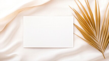 Poster - A blank rectangular card lies on soft fabric alongside a dried golden leaf, creating a serene and minimalistic aesthetic.