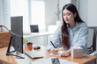 © MINAE - Young Asian businesswoman writing notes at her modern office desk with a computer, a gift, a smartphone and a takeaway coffee, she is working on a new project