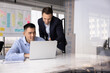 © fizkes - Male Caucasian project leader giving advice, help, instructions to Asian employee. Two colleagues talking at computer, sitting and standing at workplace, pointing and looking at screen