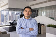 © fizkes - Confident successful young Asian company CEO man posing for business portrait, looking at camera with hands folded in confidence gesture, standing in modern office workspace alone