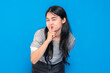 © Saddha Visual - Asian young woman with long black hair, wearing a gray shirt, playfully making a shushing gesture with her finger on lips, smiling mysteriously against a blue background.