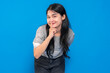 © Saddha Visual - Asian young woman with long black hair, wearing a gray shirt, playfully making a shushing gesture with her finger on lips, smiling mysteriously against a blue background.
