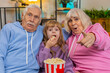 © Andrii Iemelianenko - Happy Caucasian grandfather, grandmother and granddaughter eating popcorn and watching movie on sofa at home. Smiling small girl with grandparents enjoying film during weekend in living room apartment