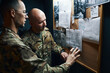 © DragonImages - Two military personnel examining and discussing strategic plans on bulletin board inside a dimly lit room, each pointing at different sections on the papers during the briefing