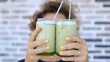 © Sia - Caucasian woman with curly hair and yellow nail polish holding two glasses of iced matcha latte with metal straws against a brick wall background