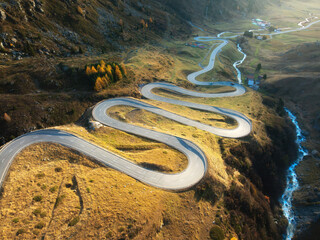 Wall Mural - Aerial view of winding mountain road, river in alpine valley at sunset in autumn. Top view of road in swiss mountain pass, yellow grass, trees and sunlight in fall. Colorful landscape. Switzerland