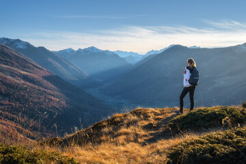 Wall Mural - Young woman with backpack standing on grassy mountain ridge enjoying inspiring view of valley with high mountain peaks during golden hour sunset in autumn. Hiking in Italy, Livigno. Girl in Alps