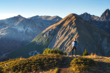 Wall Mural - Young woman with backpack standing on grassy mountain trail enjoying valley with high mountain peaks during golden hour sunset in autumn. Hiking and trekking in Italy, Livigno. Girl in Alps in fall