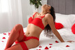 © Pixel-Shot - Beautiful young African-American woman in red lingerie with rose petals sitting on bed at home. Valentine's Day celebration