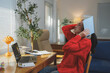 © crizzystudio - Young woman in a red sweater feeling overwhelmed while working from home. She holds a notebook over her head, sitting at a desk with a laptop, surrounded by a cozy, well-lit workspace