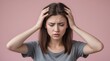© Svetlana - young woman with brown hair wearing a gray shirt. She is holding her hands on both sides of her head, expressing discomfort, stress, or possibly a headache. The background is a solid light pink color
