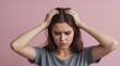 © Svetlana - young woman with brown hair wearing a gray shirt. She is holding her hands on both sides of her head, expressing discomfort, stress, or possibly a headache. The background is a solid light pink color