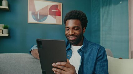 Cheerful African American man reclining on comfortable sofa holding tablet. Browsing content or watching videos while smiling. Enjoying moment at his apartment. Looking at big touch screen.