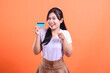 © Ajab - A cheerful young woman holds a credit card with a smile on her face and points forward, isolated on orange background