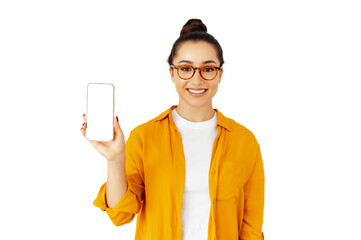 Smiling caucasian woman in a yellow shirt and glasses holds up a smartphone with a blank white mock-up screen, standing against an isolated white background. Concept of mobile advertising, technology