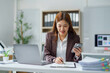 © Crystal - Young businesswoman comparing information on smartphone and financial documents while working at office desk