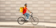 © Prostock-studio - Road to client. Young man in safety helmet with big yellow backpack goes on bicycle on gray brick wall background, outside, copy space