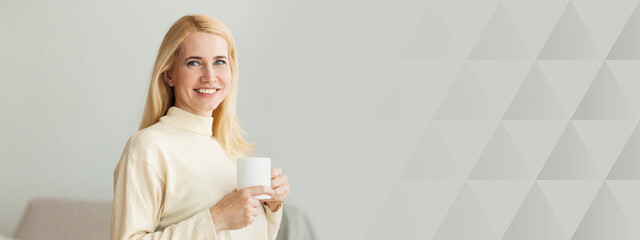 Poster - A young woman with long blonde hair enjoys a warm beverage while standing in a stylish indoor environment. Soft natural light fills the room, creating a cozy atmosphere, copy space