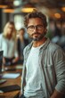 © Wilker - Close-up portrait of a man with a beard and mustache. He is wearing a grey, open, casual shirt over a light-grey t-shirt. He wears sunglasses. The background is blurred, showing indistinct figures,