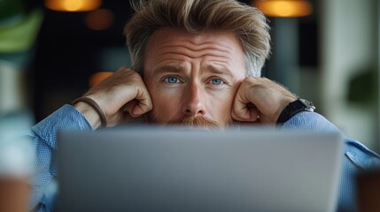 Poster - Man with a beard and blue shirt is looking at a laptop. He is wearing a watch and has his hands on his head