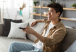 © Prostock-studio - Leisure And Entertainment. Smiling asian guy watching TV show or film, holding remote control. Young man enjoying free time sitting on the comfortable couch at home in living room, free space