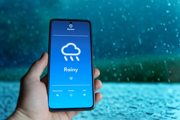 hand holding a smartphone displaying a weather app with a rainy forecast. raindrops falling in the b