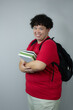 © AnaRosa - Smiling young man with curly hair wearing a red t-shirt and a backpack, holding books in his arms. Happy and friendly student portrait in studio. Education and learning concept. Copy space available.