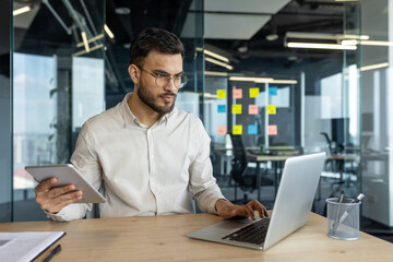 Multitasking, man with tablet computer working inside office using laptop. Businessman serious and focused typing on keyboard. Company employee thinking and concentrated.