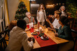© Drobot Dean - A group of multiethnic adult people of various ages sits and stands at a wooden table with food, champagne flutes, and candles in a kitchen with a Christmas tree, preparing to eat and celebrate.