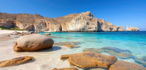 Wall Mural - crystal clear turquoise water laps smooth rocks on a pristine beach; dramatic cliffs rise in the dis