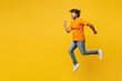 © ViDi Studio - Full body side profile view young happy Indian man he wear orange t-shirt casual clothes jump high run fast hurry up be in rush isolated on plain yellow background studio portrait. Lifestyle concept.