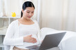 © Ekkasit A Siam - Asian pregnant woman resting on a sofa while using a laptop, touching belly with a gentle smile. Wearing a white cardigan and soft outfit, enjoying a peaceful moment in a home setting.