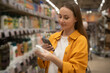 © Grustock - A shopper browses the store shelves, carefully scanning liquid soap products. She selects items while assessing labels for scent and ingredient details. The atmosphere is lively.