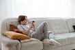 © Kokhan O - Teenager works on homework while relaxing on a couch in a cozy living room during the afternoon. Full length portrait of young teenage boy in hand smartphone while sitting on sofa