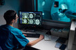 © pressmaster - Medical professional reviewing patient's MRI images on computer monitor with patient in MRI machine in background. Hospital equipment and technology setup visible