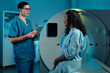 © pressmaster - Medical professional discussing diagnosis with patient seated beside an imaging machine. Patient wearing hospital gown while doctor holds device, creating a focused environment in medical facility
