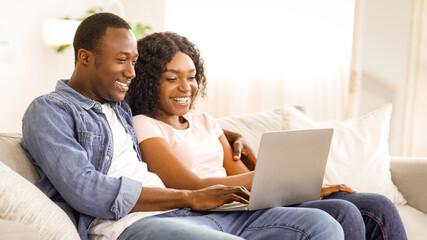 Cheerful black couple using laptop while resting on couch at living room at home, watching funny content together, copy space