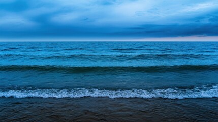 Wall Mural - Calm ocean waves gently lap against a sandy shore as dusk approaches, with dark clouds creating an atmospheric twilight backdrop over the water's surface