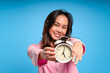 © Daniel - Asian woman in pink pajamas smiling warmly while holding a classic white alarm clock toward the camera, against a solid blue background, symbolizing morning routine and wake-up time