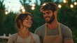 © fotogurme - Smiling couple in aprons outdoors, rustic celebration, romantic evening, festive string lights, Euro-American