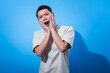 © Daniel - Asian man expressing shock or surprise with both hands on his cheeks and mouth open. Wearing a white t-shirt and posing dramatically against a solid blue background
