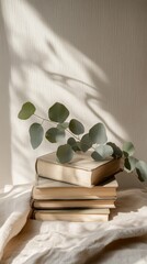 Poster - A stack of old books rests on a light fabric, adorned with fresh eucalyptus sprigs. Gentle sunlight creates soft shadows, evoking a peaceful atmosphere perfect for relaxation and reading