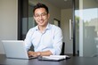 © SimpleDesignStudio - Smiling young Asian male professional working on a laptop in a modern office environment, showcasing a confident and engaged attitude towards his tasks.