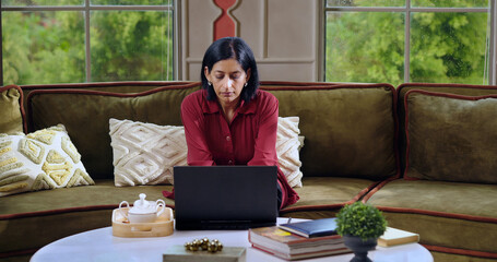 Indian mature gen x busy woman lady sitting on sofa couch use laptop look web screen do online work from home check new study email read casual news female spend day time indoor house