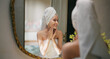 © Raushan_films - Indian young adult gen z woman lady wrapped towel on head and body look mirror apply make up cream on dry cheek face teen age girl female hold bottle take liquid hand do fresh day skin care house home