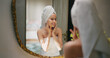 © Raushan_films - Indian young adult gen z woman lady wrapped towel on head and body look mirror apply fresh make up cream on dry cheek face teen age girl do female hand facial massage day skin care house home
