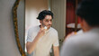 © Raushan_films - Indian Asian gen z young adult smiling guy bearded man look mirror hold soft brush apply white foam gel cream to his beard face before shave do morning day self skin care routine indoor house
